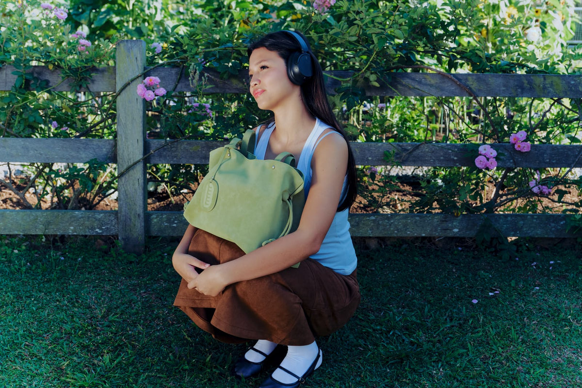Isabelle Bag in Meadow — campaign, crouching by a rose-covered fence with headphones